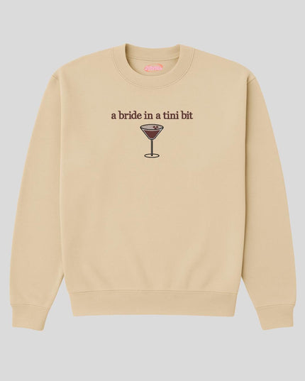 A beige sweatshirt with a graphic design depicting a glass of wine and the text "a bride in a tini bit" printed on it.