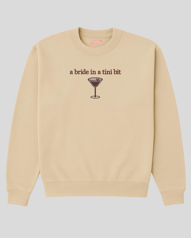 A beige sweatshirt with a graphic design depicting a glass of wine and the text "a bride in a tini bit" printed on it.