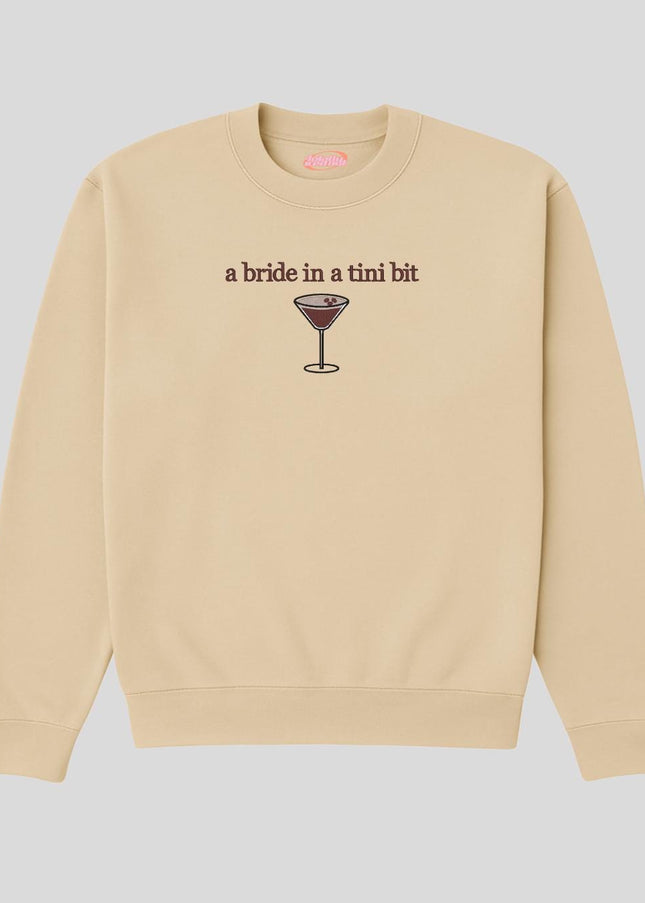 A beige sweatshirt with a graphic design depicting a glass of wine and the text "a bride in a tini bit" printed on it.